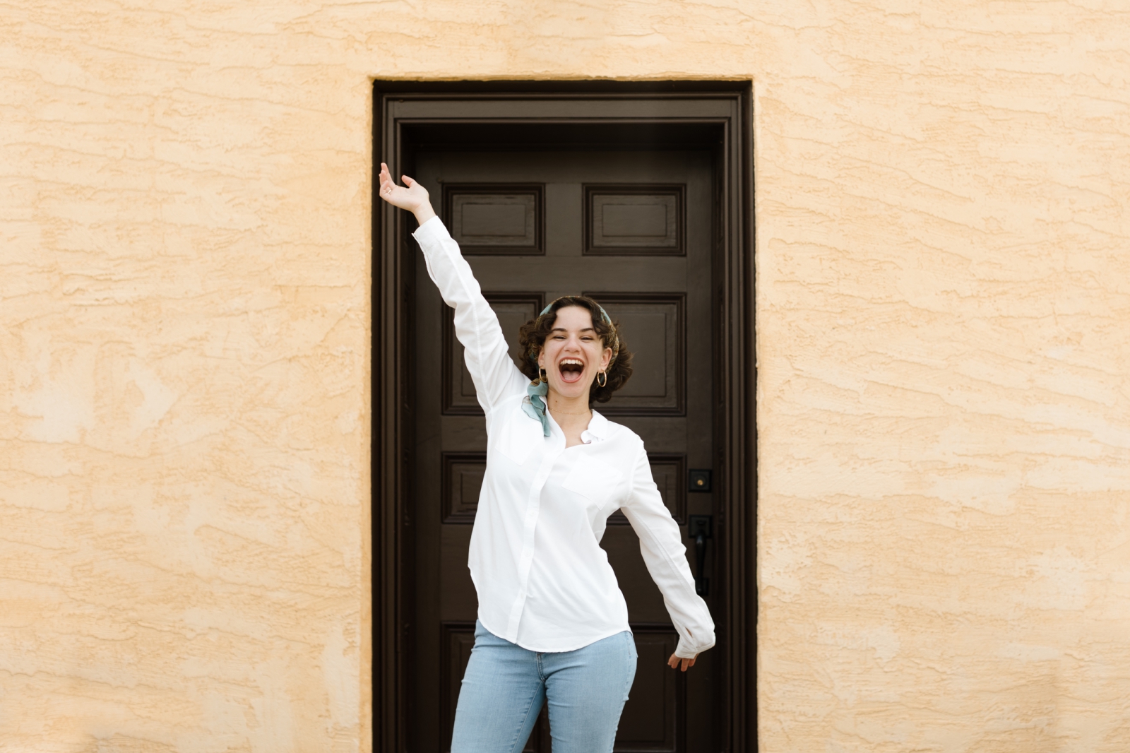 A headshot of a website copywriter standing with arms outstretched in front of a brown door set into a yellow wall