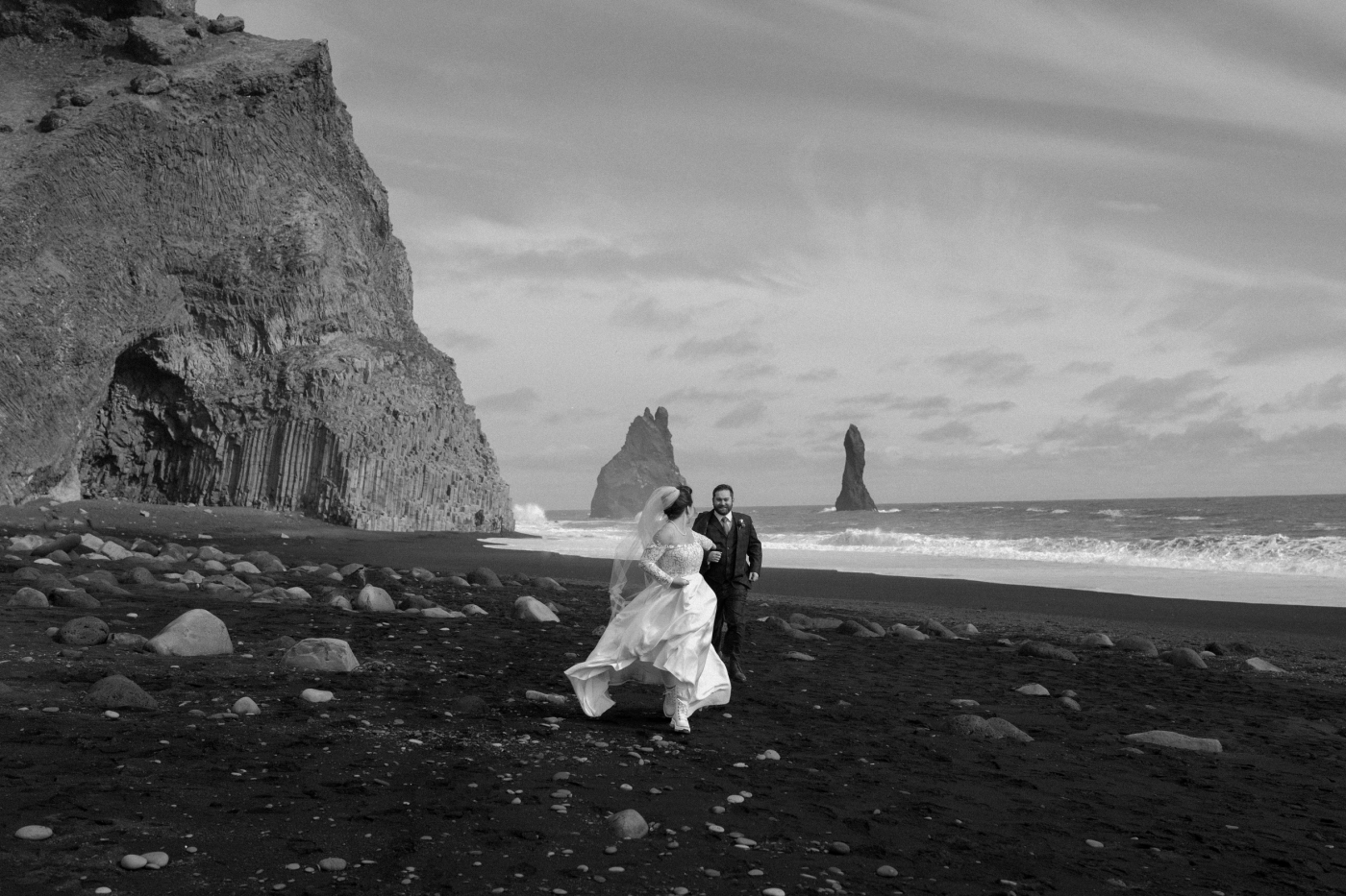 A black and white photo of a bride and groom running on Reynisfjara black sand beach
