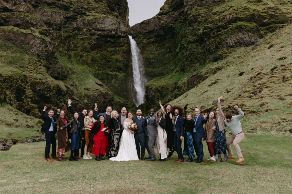 A group of twenty wedding guests surround the bride and groom and stand in front of a waterfall